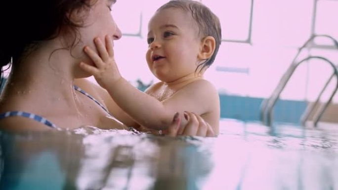 妈妈和快乐的宝宝在室内游泳池里抚摸着微笑的脸
