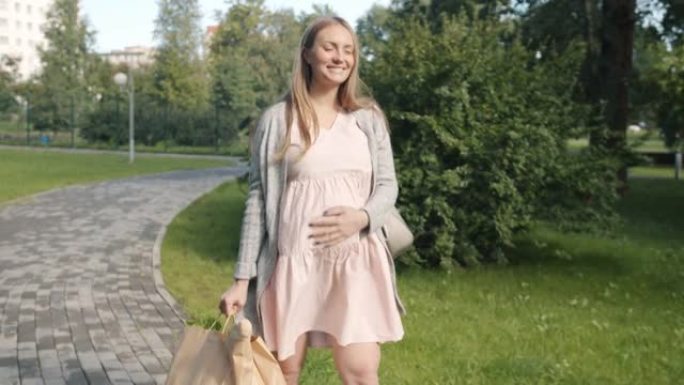 多莉拍摄的快乐孕妇走在城市公园里，带着健康食品的纸袋