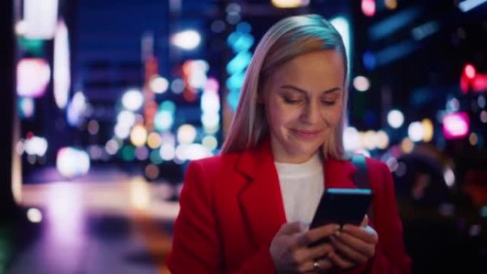 一个穿着红色外套的美女的肖像在夜晚霓虹灯的现代城市街道上行走。迷人的女性使用智能手机，环顾城市电影环