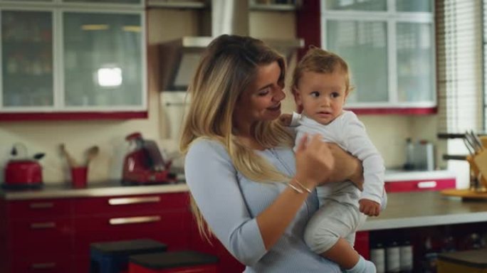 一位年轻快乐的微笑母亲的真实镜头将她蹒跚学步的男婴抱在怀里，并在家里的厨房里一起跳舞。
