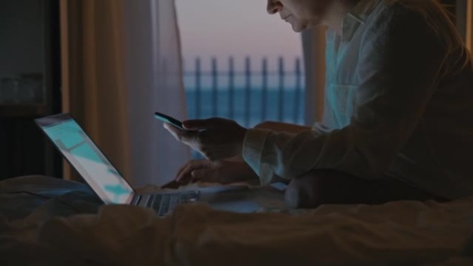 女人晚上坐在旅馆房间的床上，在笔记本电脑中从智能手机中输入信息