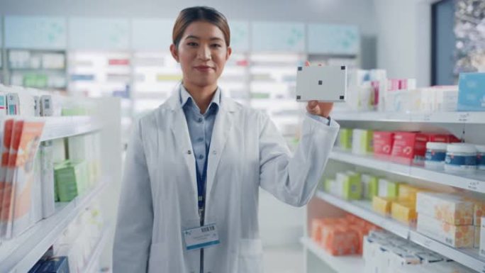药房药店: 专业亚洲女药剂师的肖像拿着带有跟踪标记的模拟模板药包，看着相机，微笑着。专家推荐最佳产品