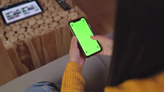 年轻女士无法辨认的女人坐在家里的沙发上时使用带有色度键屏幕的智能手机