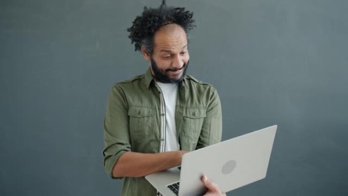 快乐的阿拉伯男人的肖像使用笔记本电脑，然后看着相机，然后在灰色背景上眨眼
