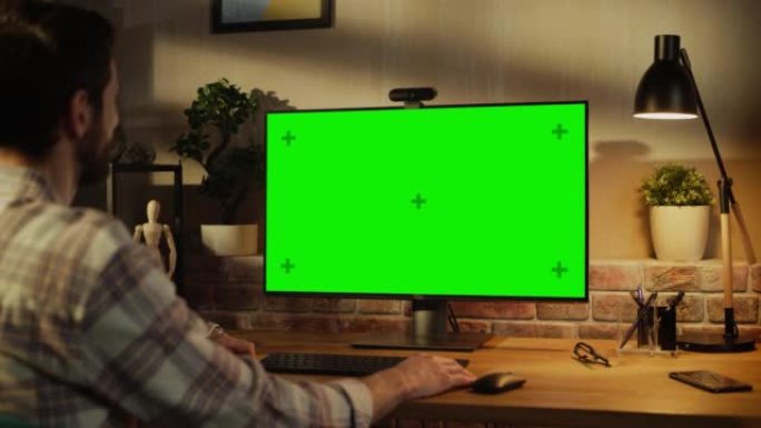 办公室员工在家工作，在带有绿屏模拟显示器的台式计算机上进行视频通话。男人坐在客厅里，晚上与朋友和家人