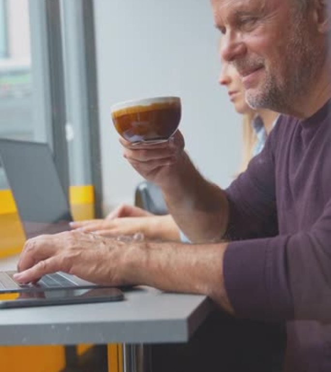 顾客在咖啡店橱窗里在笔记本电脑上工作和在手机上浏览的垂直视频