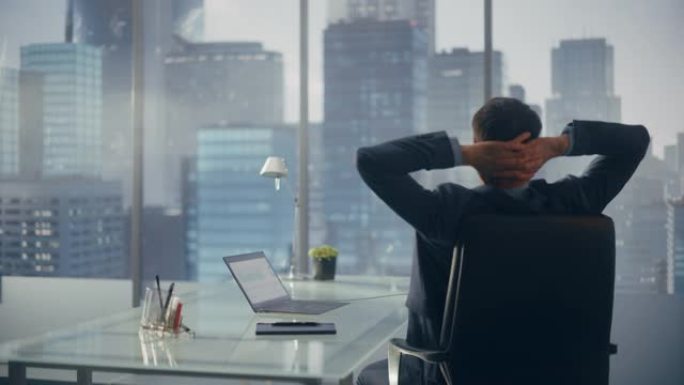 穿着西装的轻松商人坐在现代办公室的办公桌前，使用笔记本电脑，看着窗户的城市景观。成功的财务经理在计划