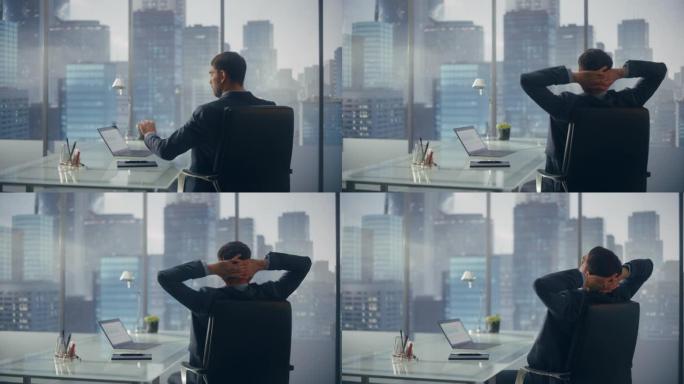 穿着西装的轻松商人坐在现代办公室的办公桌前，使用笔记本电脑，看着窗户的城市景观。成功的财务经理在计划