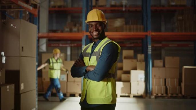 英俊快乐的职业工人穿着安全背心，戴着安全帽，微笑着在镜头前交叉双臂。在后台的大仓库里，货架上堆满了送