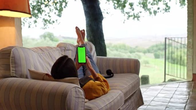 DS年轻女子躺在露台上的沙发上，使用带色度键绿屏的智能手机