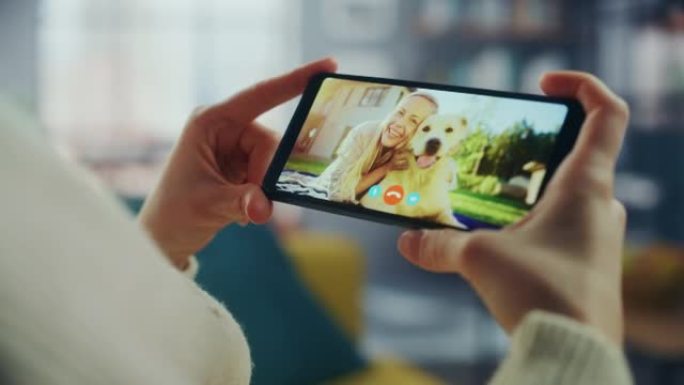 一位女性在视频通话中与她美丽的朋友在家庭客厅的智能手机上与狗聊天的特写镜头。自由职业者通过互联网与客