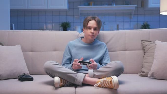 年轻英俊的游戏男孩坐在沙发上，在数字控制台上玩视频游戏的肖像。兴奋的玩家控制视频游戏操纵杆。一个人专