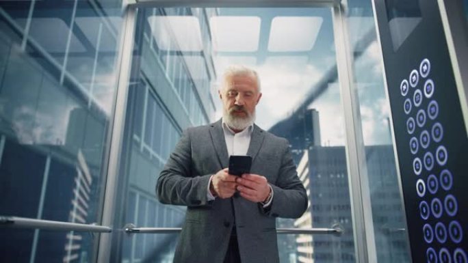 成功的中年商人在现代商务中心乘坐玻璃电梯到办公室。英俊的快乐男人使用智能手机，在电梯里写信息、社交媒