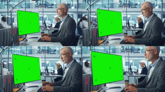 高加索高级专家在繁忙的公司办公室中使用带有绿色色度键屏幕显示的台式计算机。成熟的男性经理创造性地在多