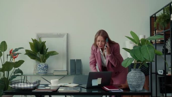 在办公室工作的快乐成功美丽的女商人通过手机聊天，使用笔记本电脑咨询客户。