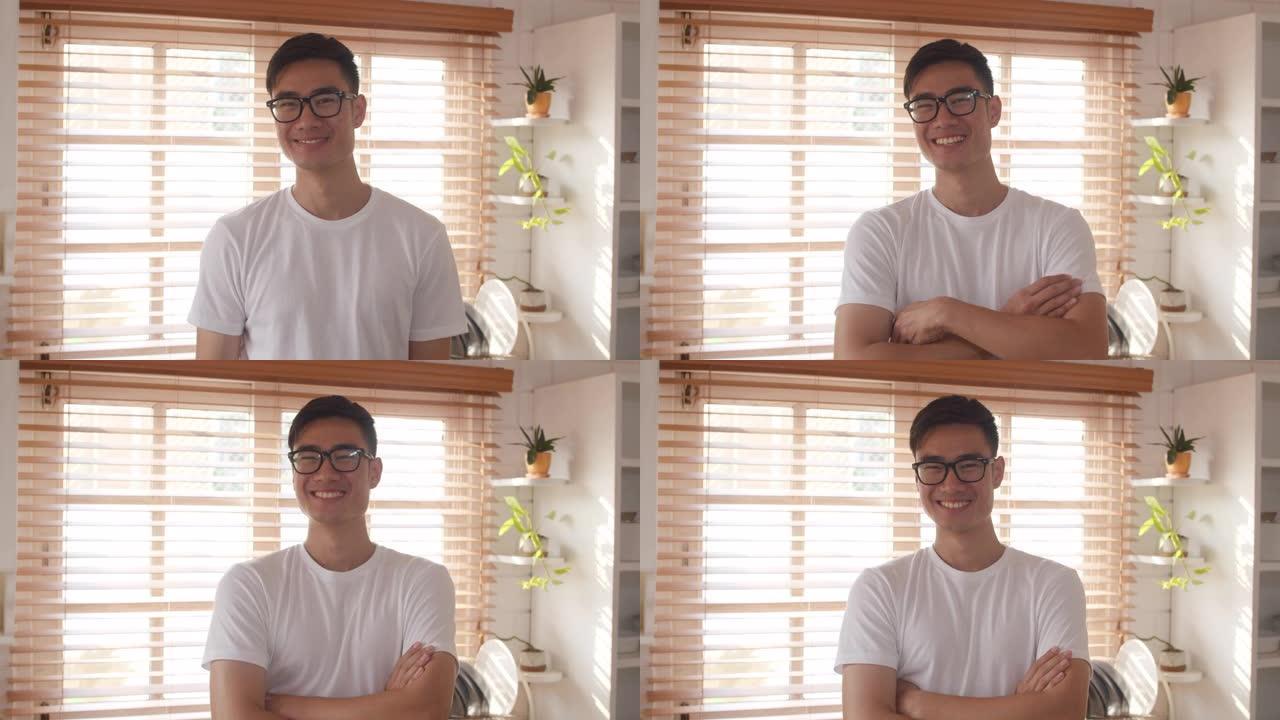 快乐的年轻亚洲男性在家里的厨房里放松时，微笑着快乐，双臂交叉，看着镜头。