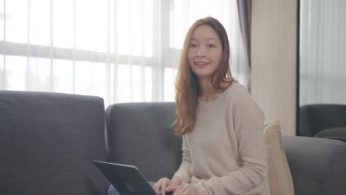 美丽的亚洲年轻女子年龄36岁在家里工作的笔记本电脑看着相机，而坐在沙发上放松早上在家。成人在线学习概