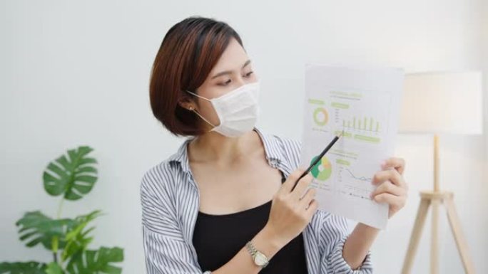 亚洲女商人在预防病毒的情况下戴着口罩社交距离，看着摄像机向同事介绍办公室视频通话工作中的计划。