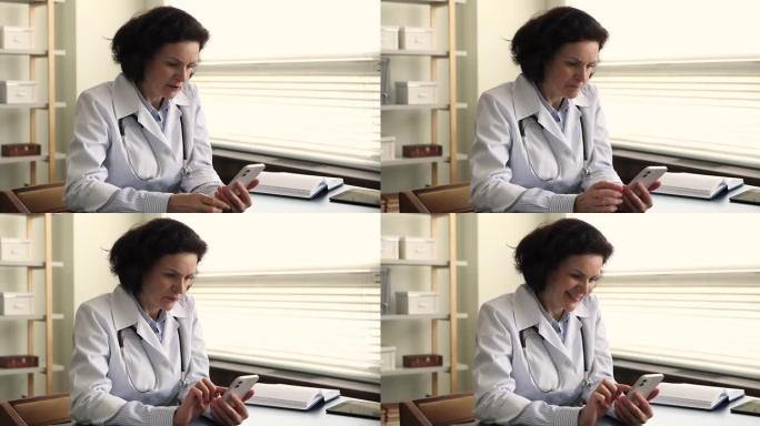 穿着外套的成熟女医师坐在办公桌前拿着手机