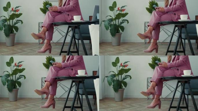 特写优雅的商务女性穿着粉色西装，使用智能手机应用程序，咖啡坐腿交叉在办公室桌子上。