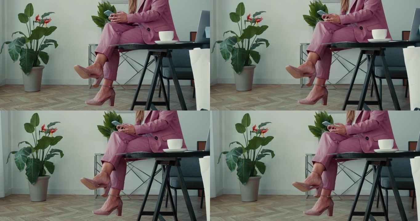 特写优雅的商务女性穿着粉色西装，使用智能手机应用程序，咖啡坐腿交叉在办公室桌子上。