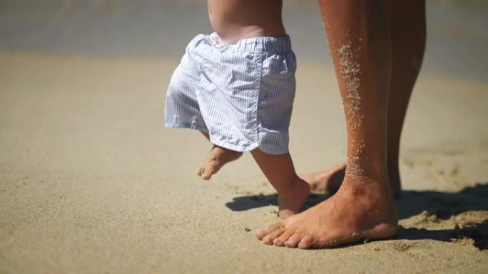 一位年轻母亲的真实特写镜头正在帮助她蹒跚学步的男婴迈出第一步，并教她在沙滩上散步