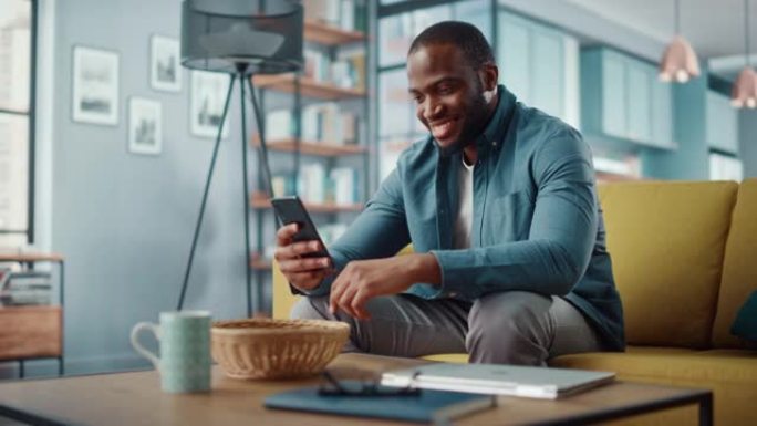 英俊的黑人非洲裔美国男子坐在舒适的客厅的沙发上使用智能手机。自由职业者在家工作。浏览互联网，使用社交