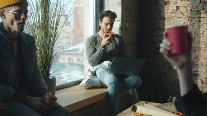 在办公室里吃披萨的商人在白天聊天和使用笔记本电脑