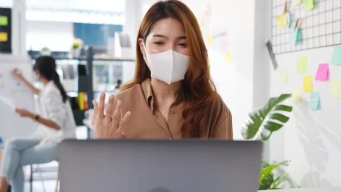 亚洲商界女性在新常态下戴着口罩进行社交距离以预防病毒，同时在办公室用笔记本电脑视频通话向同事介绍计划