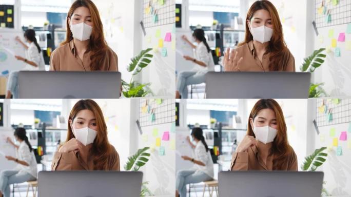 亚洲商界女性在新常态下戴着口罩进行社交距离以预防病毒，同时在办公室用笔记本电脑视频通话向同事介绍计划