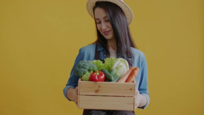 穿着制服拿着木箱的女农民肖像，黄色背景上放着新鲜蔬菜