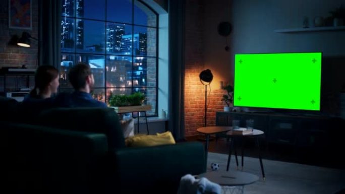 年轻的夫妇花时间在家里，坐在沙发上，在他们时尚的阁楼公寓里看绿屏模拟显示的电视。男人和女人流晚间电影