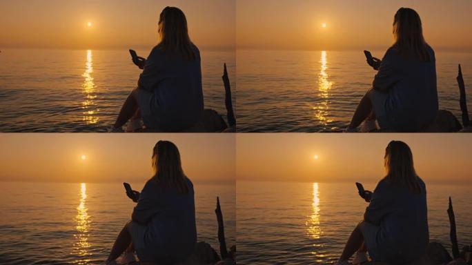 一个女人的SLO MO剪影在日落时在海岸上使用她的智能手机