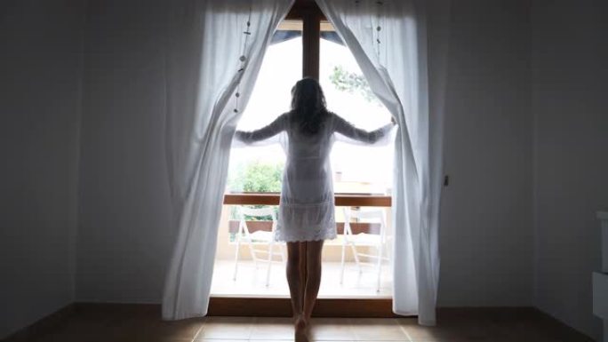 一个穿着白色连衣裙的年轻晒黑的赤脚女人的真实镜头正朝着窗户醒来，早上在酒店房间里拉开窗帘