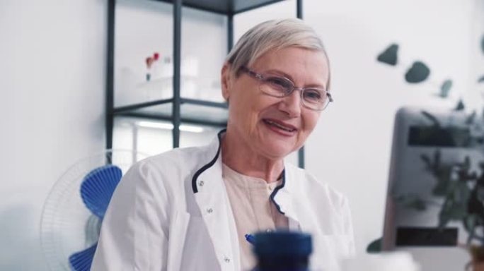 穿着实验室外套的友好积极的高级女医生微笑着在轻型办公室使用笔记本电脑网络摄像头咨询远程客户。