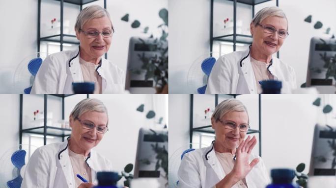 穿着实验室外套的友好积极的高级女医生微笑着在轻型办公室使用笔记本电脑网络摄像头咨询远程客户。