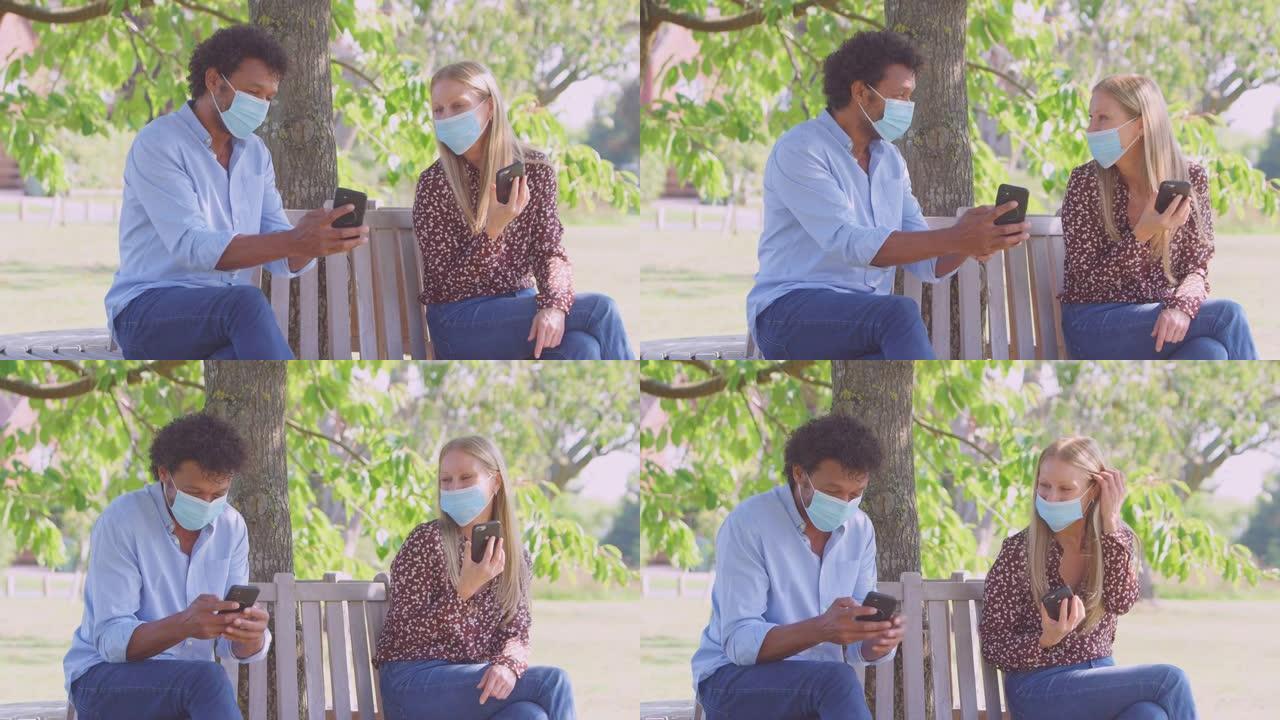 戴着口罩的社交距离夫妇在健康大流行期间看着手机在公园见面