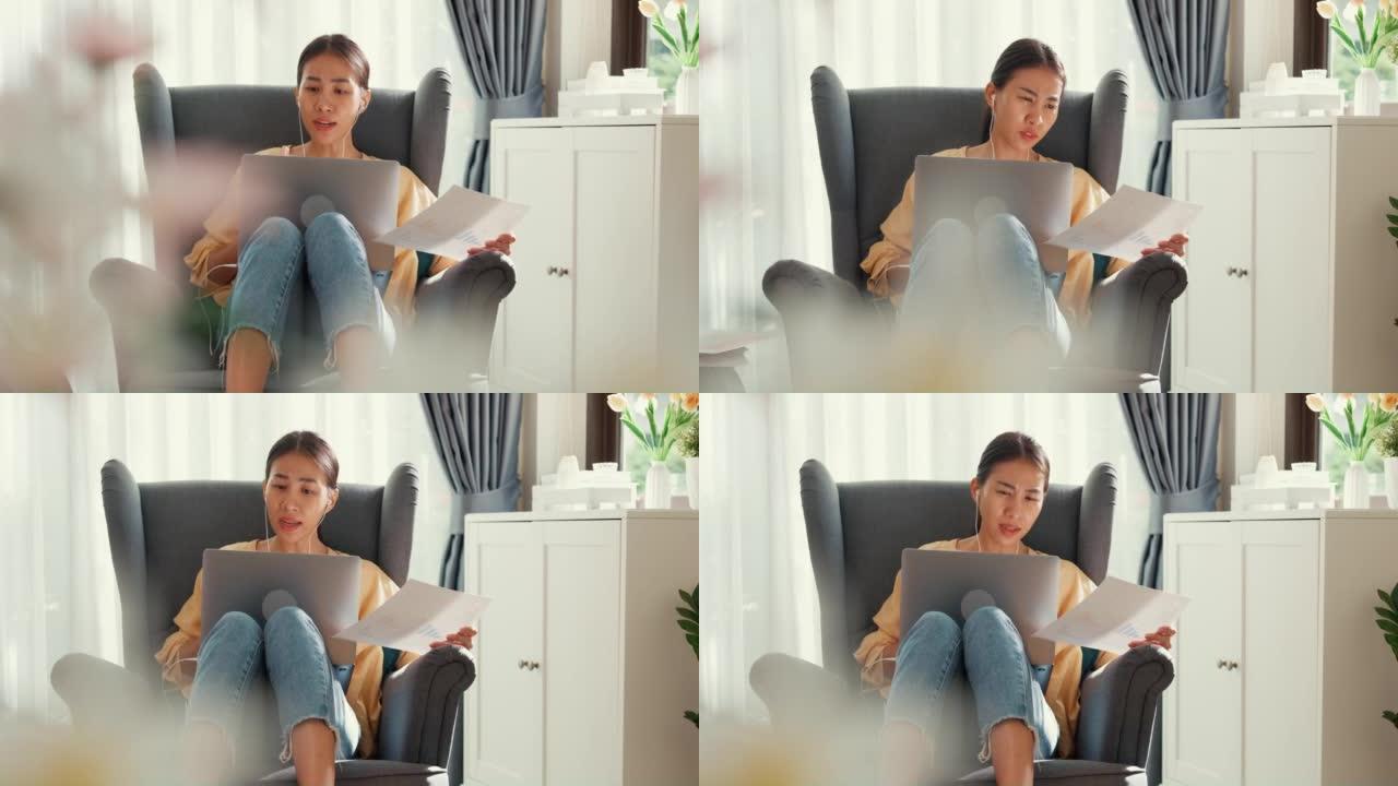 年轻的亚洲女学生或大学生坐在沙发椅上，戴着电脑笔记本电脑，戴着耳机，举行文书工作在线会议，在家里的客