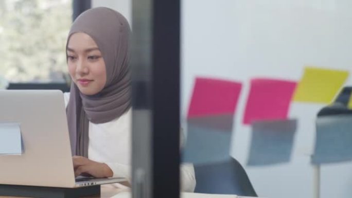 美丽的亚洲穆斯林女士休闲服在现代新的普通办公室使用笔记本电脑工作。