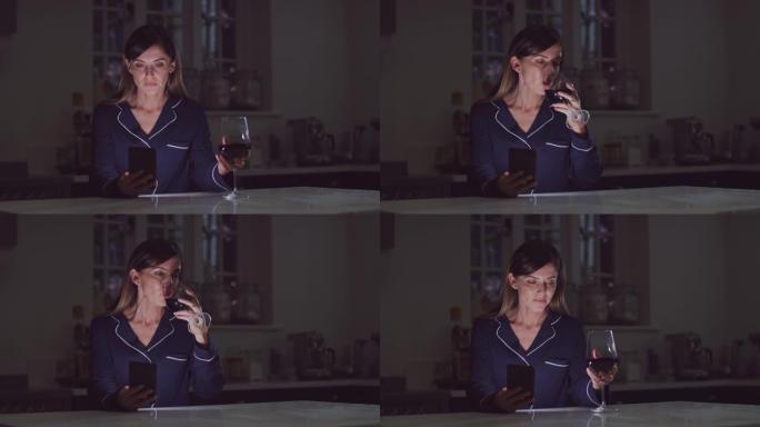 不开心的女人穿着睡衣晚上用手机坐在厨房里喝杯酒