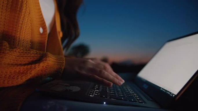 一个无法辨认的中年黑发女人坐在户外时在笔记本电脑上打字的特写慢动作，背景是黄昏的天空