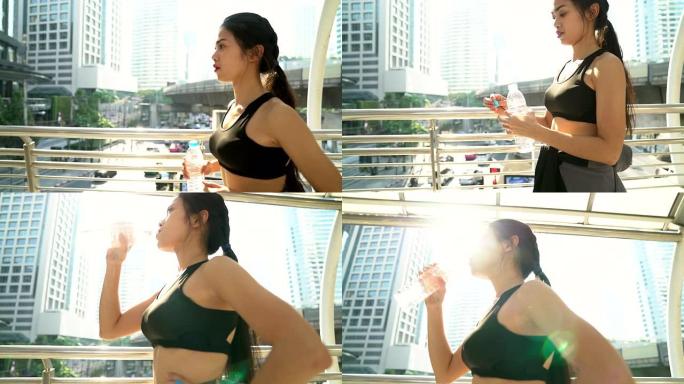 穿着健身服装的年轻女性举起水瓶喝，为跑步做准备。拍摄于早晨阳光照在她身后时拍摄的背景为高楼的城市。