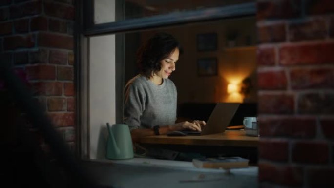 微笑的西班牙女人在温暖舒适的晚上在家使用笔记本电脑。专业自由职业者在家远程工作。从外面拍摄到公寓的窗