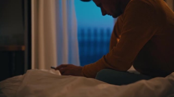 晚上，棕色短发的男人坐在旅馆房间的床上，看着他的智能手机时感到压力很大。