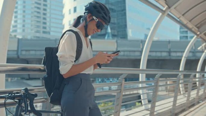 带背包的快乐亚洲女商人在城市用智能手机站在街上骑自行车去办公室上班。