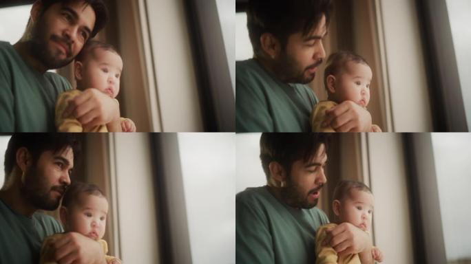 父爱的慢动作肖像: 一个年轻的父亲抱着小婴儿，他透过窗户说话和看着。男人和他可爱的蹒跚学步的孩子一起