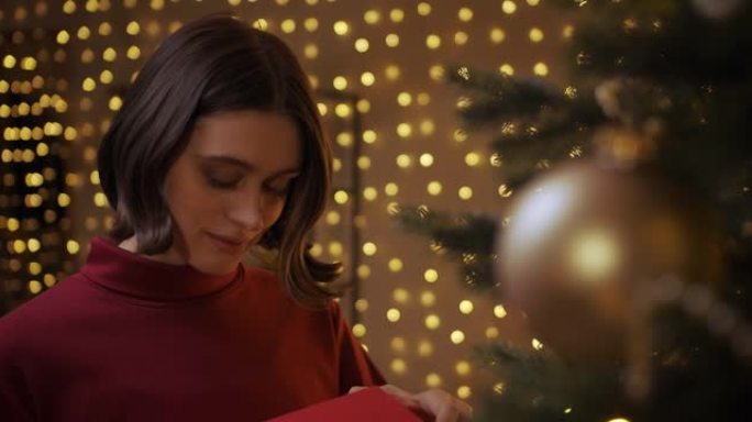 一个穿着红色毛衣的美丽微笑的女孩打开了一个要点盒，站在圣诞树附近