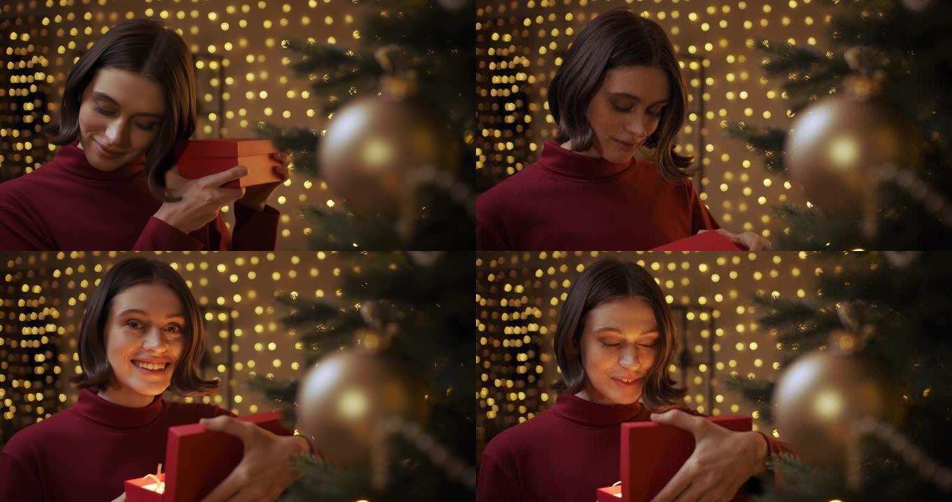一个穿着红色毛衣的美丽微笑的女孩打开了一个要点盒，站在圣诞树附近