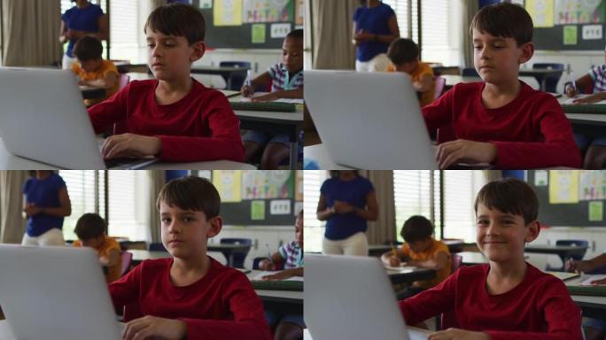 快乐的高加索男生的肖像坐在教室里，使用笔记本电脑，看着相机