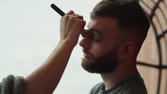 专业化妆师的特写手在美容工作室沙龙的年轻大胡子男模脸上放色调粉